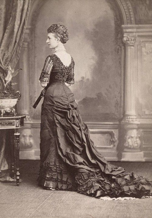 Queen Alexandra facing away from the camera, showcasing her back and the cascade of fabric bustle leading to a lengthy train, ca 1877